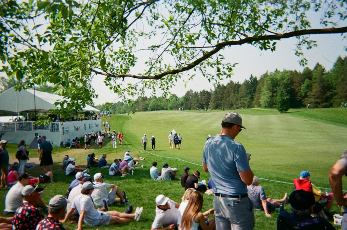 Spectators seated on the grass at a golf tournament, watching play on the fairway.