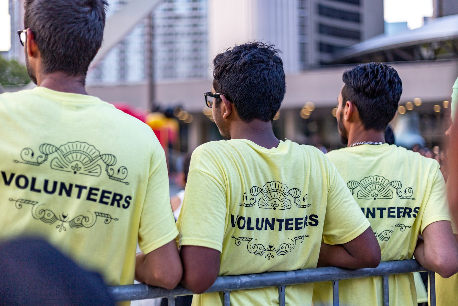 Volunteers wearing event-branded shirts at an outdoor gathering, leaning on a barrier fence.